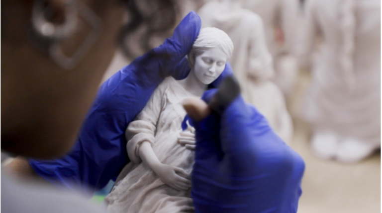 Manos con guantes azules retocan una réplica de la Madre de la Esperanza, con más figuras al fondo del taller