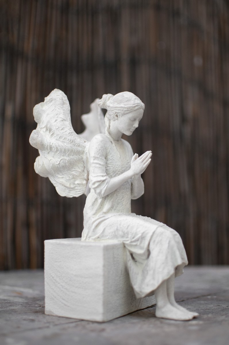 Escultura del Ángel de la Guarda sentado, con las manos juntas en oración, en el exterior.