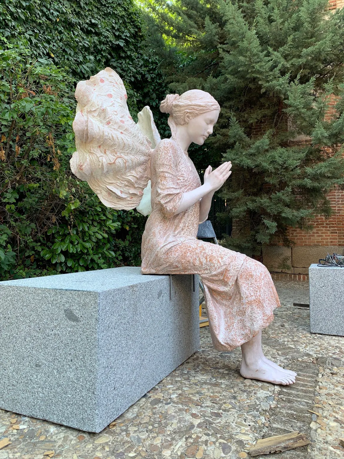 Escultura sedente de un ángel en oración sobre un pedestal de granito, en un jardín