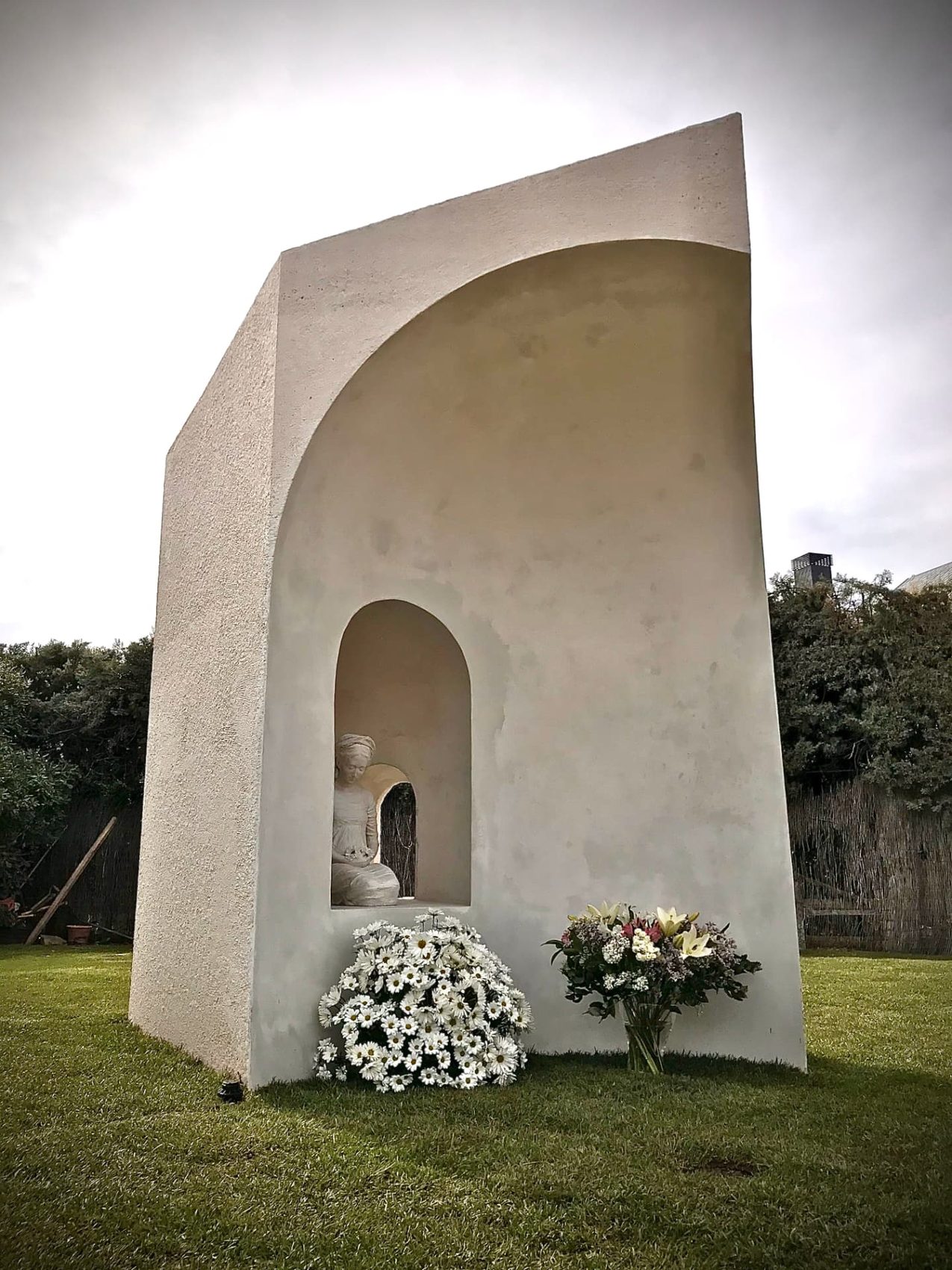 Estructura escultórica moderna de color crema en un jardín, con un nicho que contiene una estatua femenina sedente y flores a sus pies