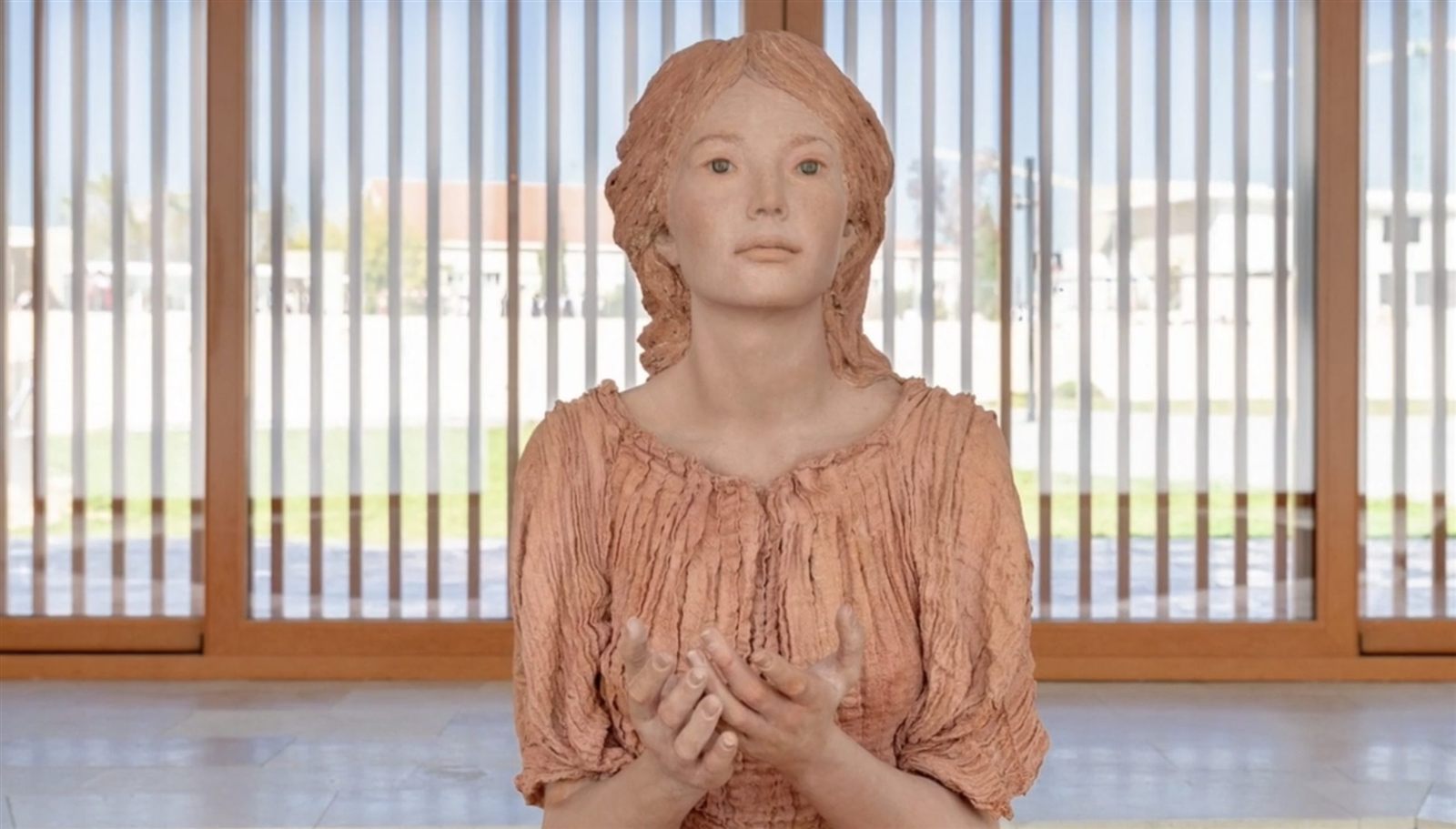 Nuestra Señora de Alegra en terracota, con las manos abiertas, ante un ventanal de lamas de madera.