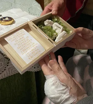 Unas manos sostienen una caja de madera abierta con la miniatura de la Virgen sobre musgo, y otra mano la recibe.