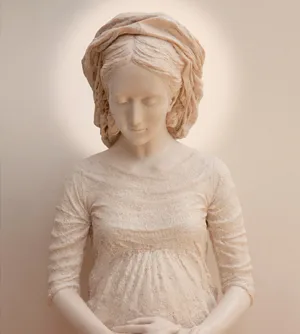 Busto de la Virgen con la mirada baja, iluminada por un halo cálido sobre la pared.