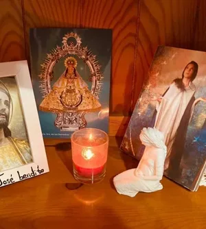 La miniatura de la Virgen junto a una vela roja encendida y varias estampas, sobre una mesa de casa.