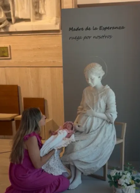 Una mujer arrodillada sostiene a su bebé recién nacida ante la escultura de la Madre de la Esperanza, en una capilla. Sobre el panel se lee «Madre de la Esperanza, ruega por nosotros».
