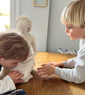 Dos niños contemplan de cerca, sobre la mesa de casa, la escultura de la Virgen sentada.