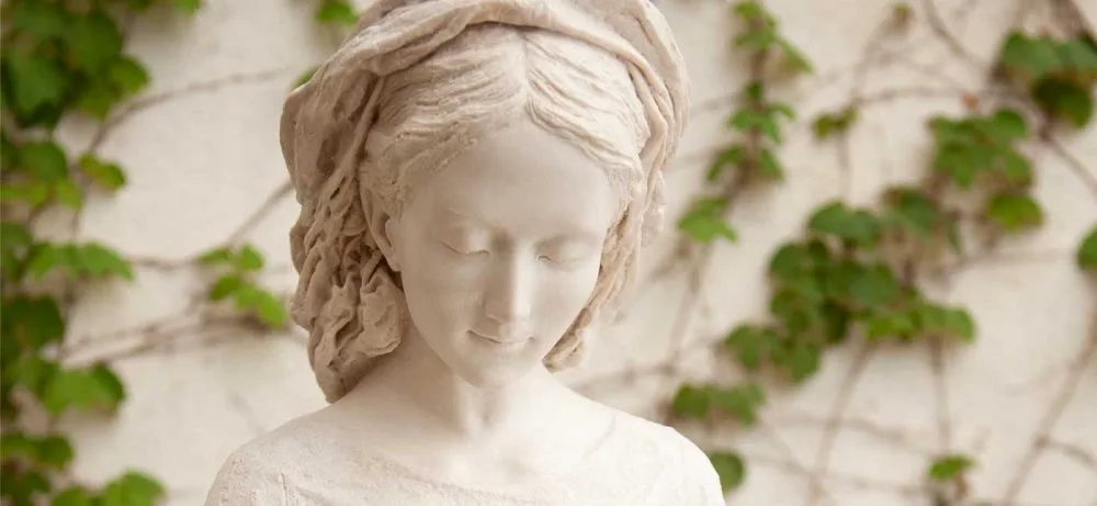 Detalle del rostro de la Virgen, con los ojos bajos, ante un muro cubierto de hiedra.