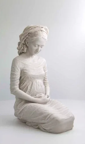 La Virgen de Hakuna, sentada y embarazada, con las manos sobre el vientre, sobre fondo neutro.